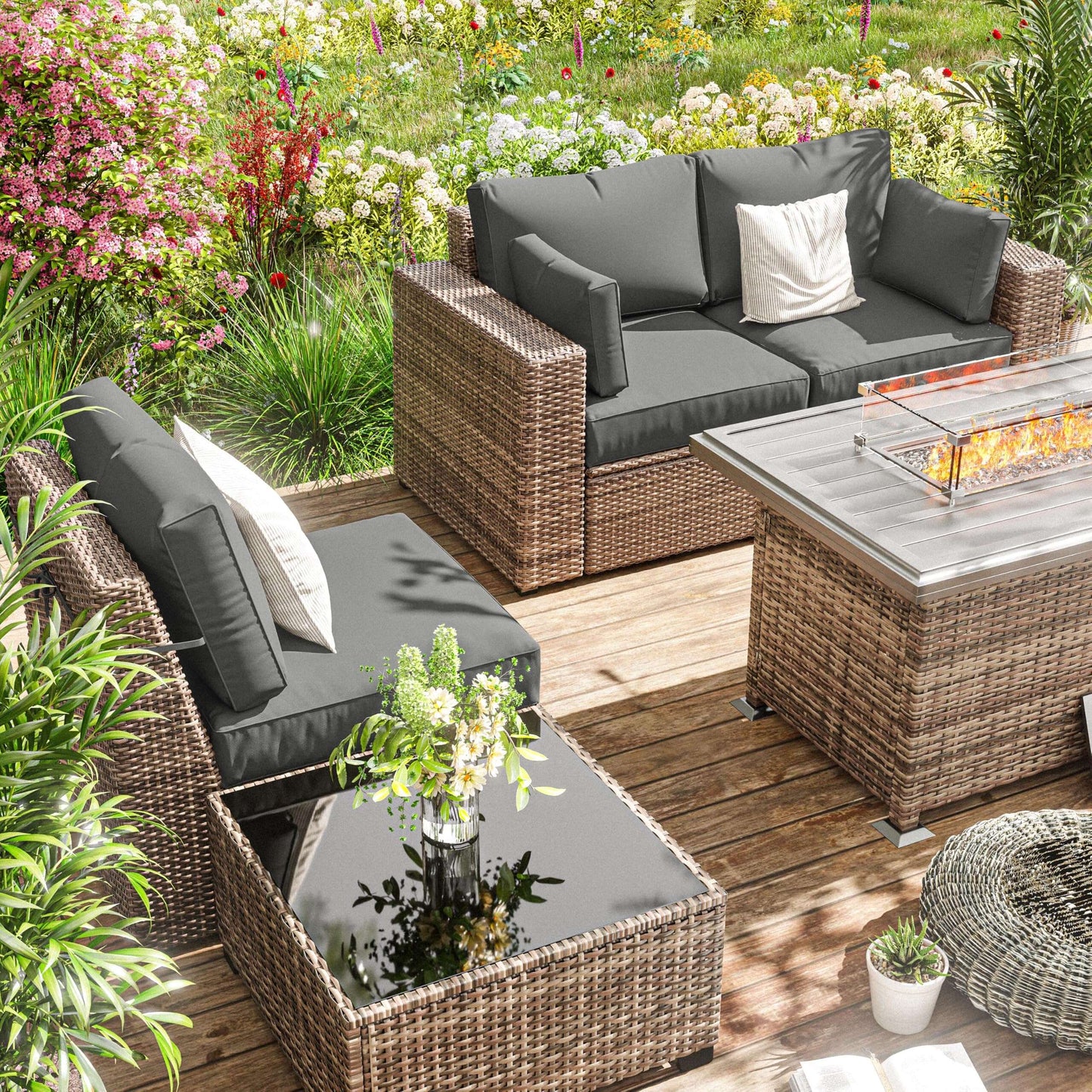 Outdoor wicker patio furniture set with grey cushions, fire pit table, and garden flowers