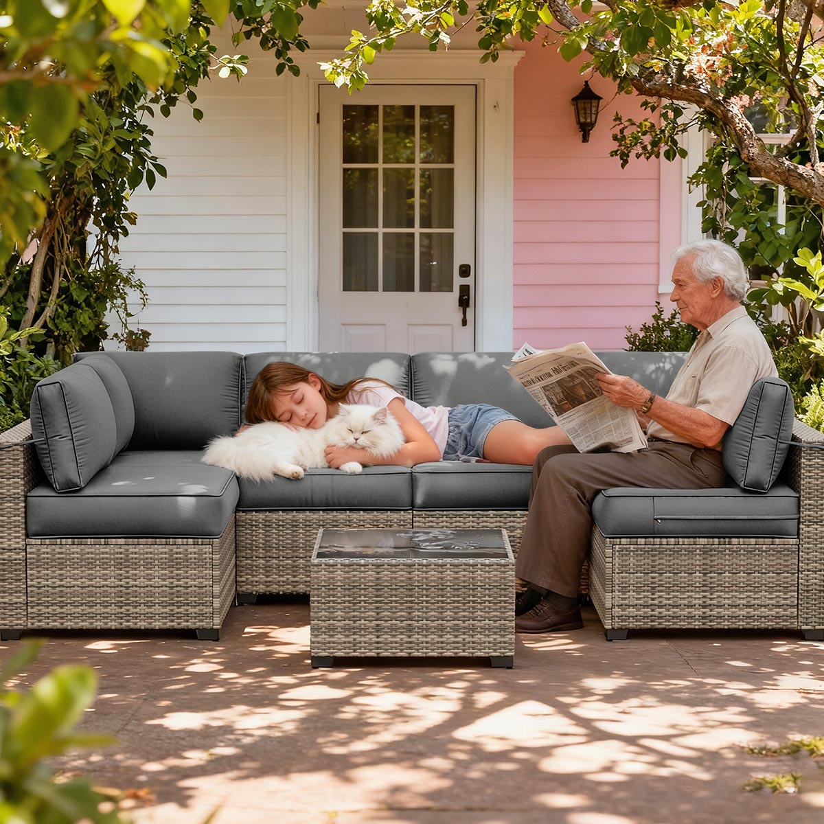 Two people sitting on a gray outdoor sofa with a white dog, reading a newspaper, in front of a pink house.